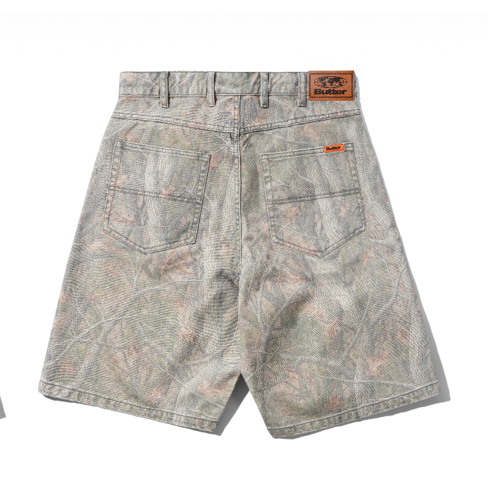 Back view of a pair of camouflage-patterned denim shorts with a brown leather patch on the waistband and orange brand labels on the back pockets, set against a plain white background.