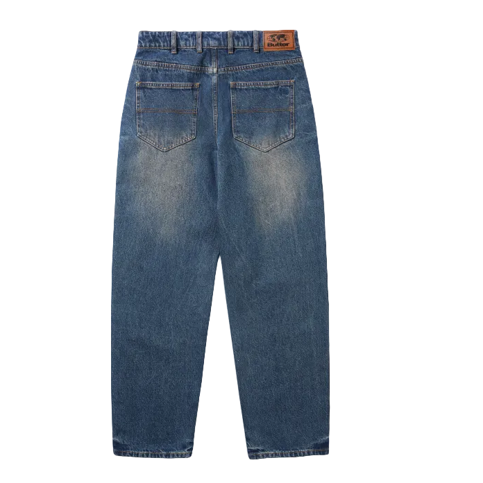 Back view of a pair of blue denim jeans with a faded wash, featuring a brown leather patch on the back waistband and two back pockets, displayed on a white background.