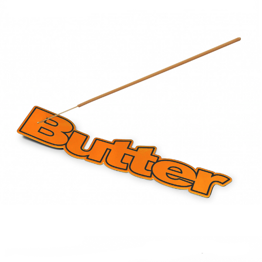 An orange metal incense holder spelling 'Butter' in 3D block letters, shown with a single incense stick resting diagonally across it on a white background. Text below reads 'Metal Incense Holder' and 'Orange' with product dimensions.